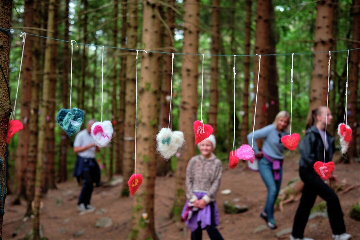 Foto som syner filthjarter som heng på ei snor i ein skog. Bak står det ei jente og smiler medan folk går forbi bak ho att.