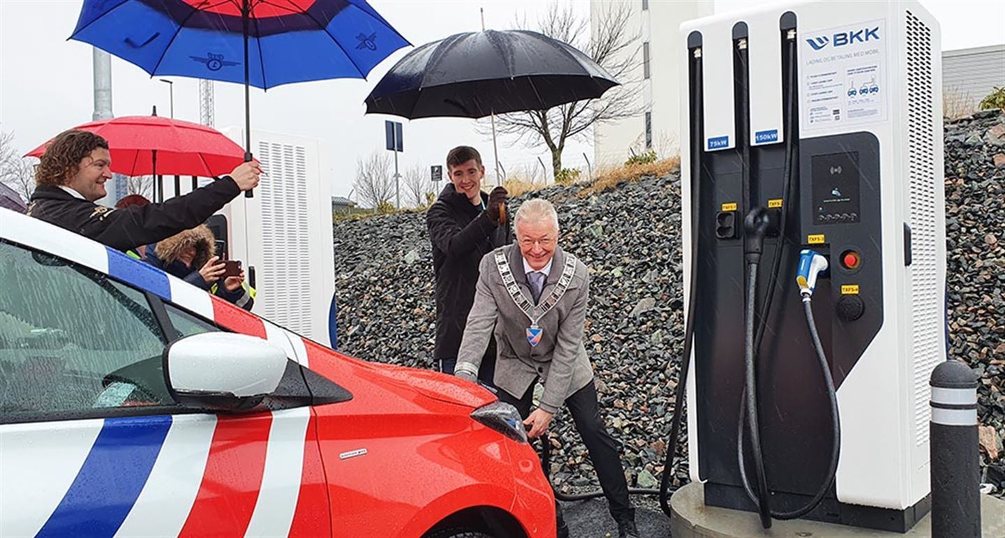 Fylkesordførar Askeland festkledd med ordførarkjede ladar ein bil. Bak han står ein mann å held paraplyen for han. Foto.