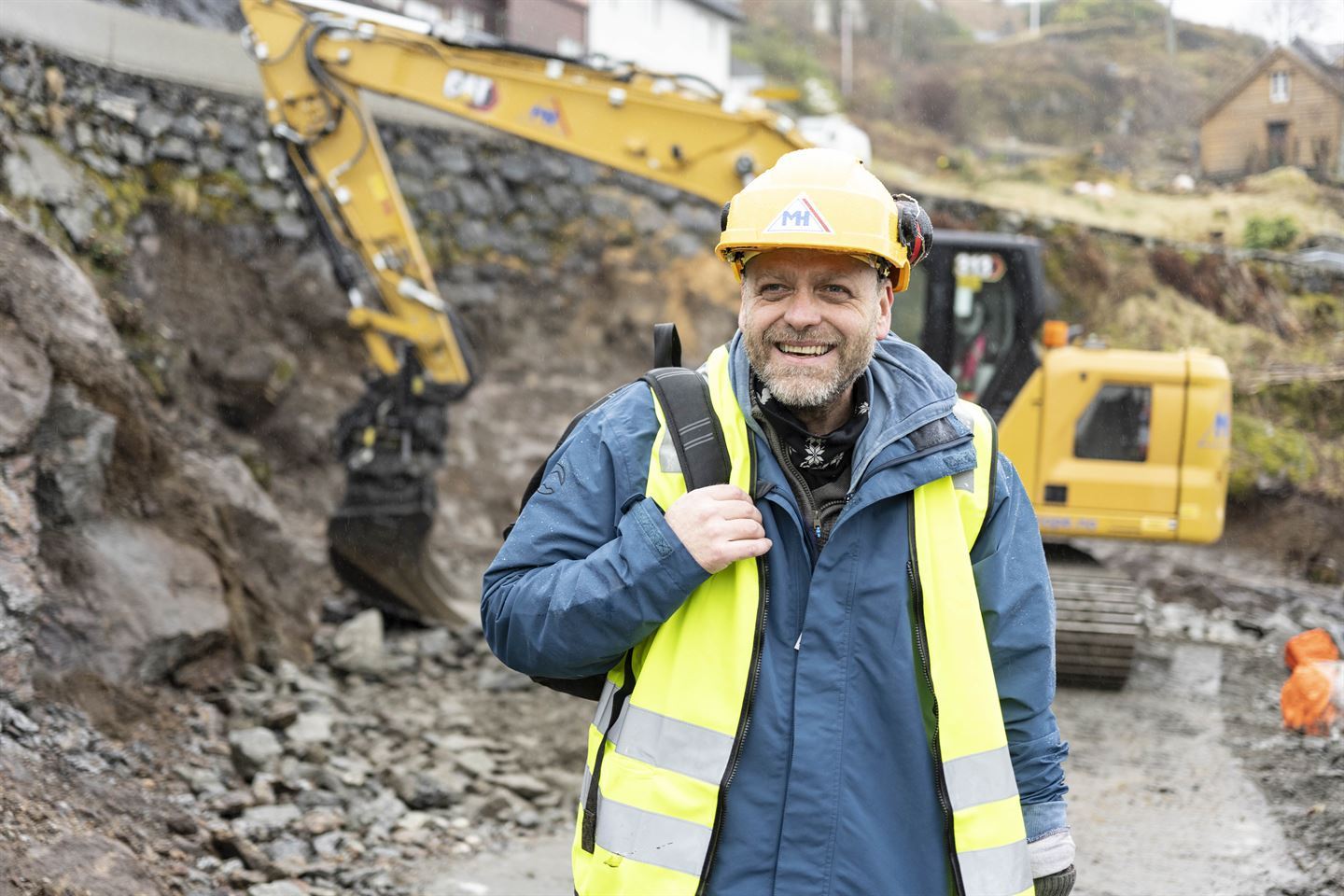 mann med vernehjelm smiler med gravemaskin i bakgrunnen