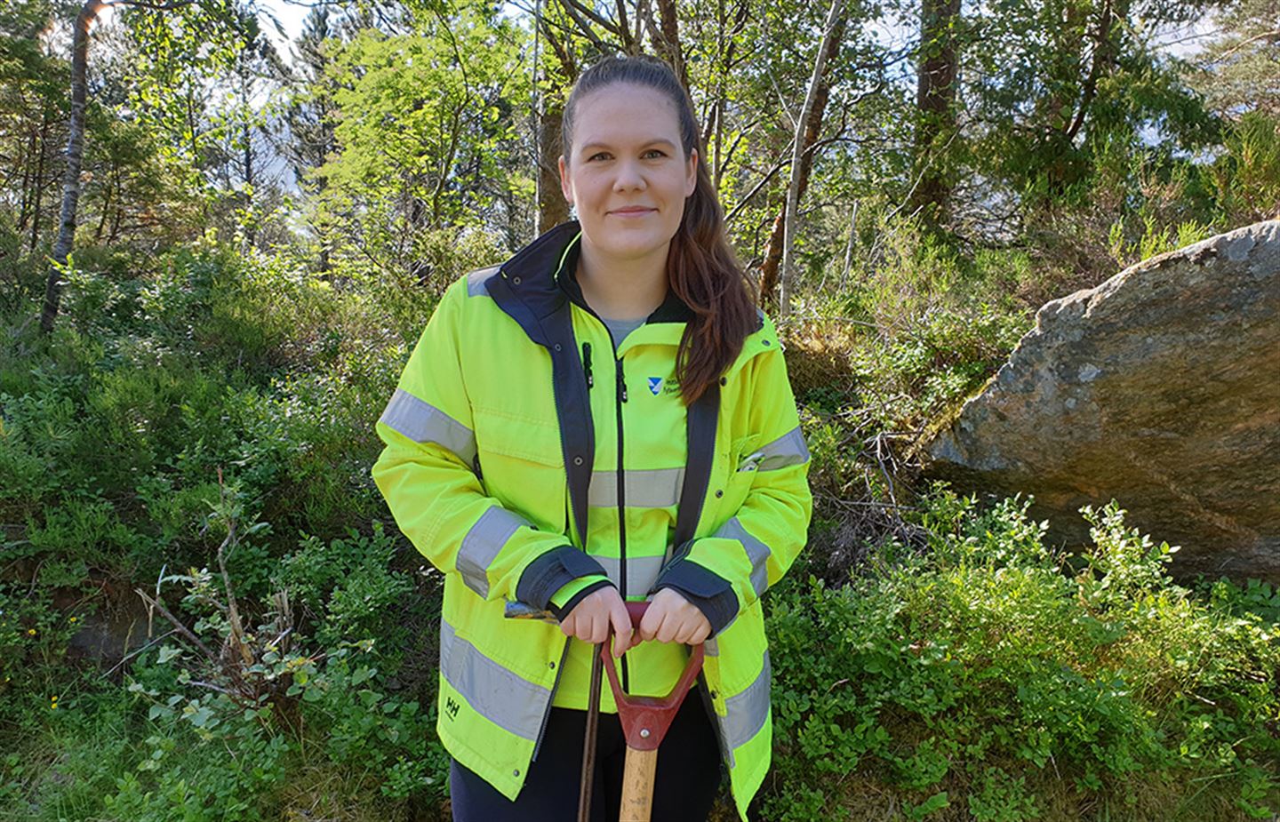 Foto av kvinne i arbeidsklede og med ein spade ute i naturen.