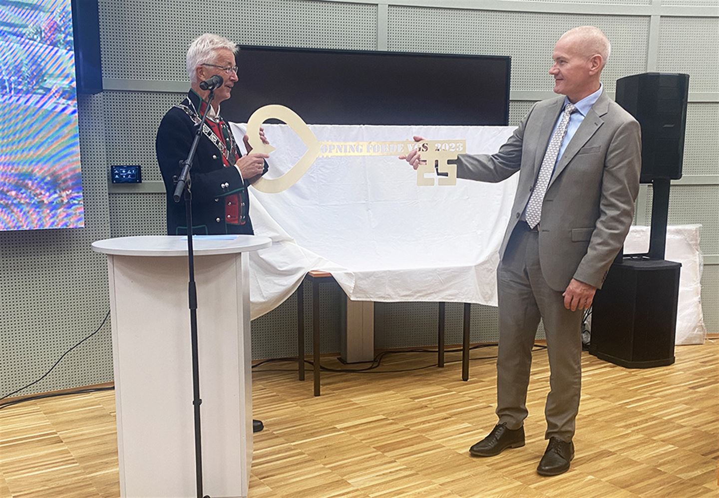 Mann i bunad og mann i dress held stor nøkkel mellom seg.
