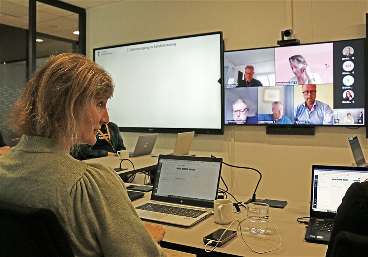 Person i rom fotografert bakfrå med skjermar med personar på veggen i bakgrunnen. 