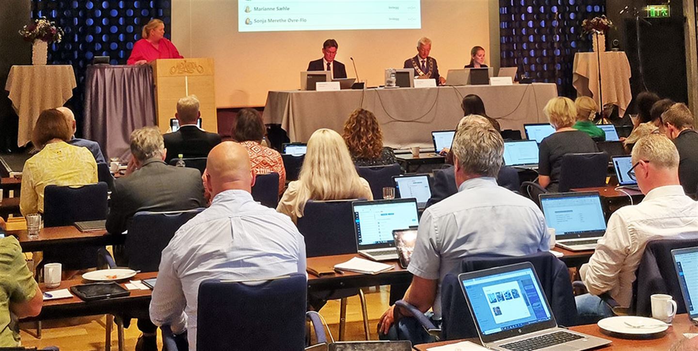 Personar i ein stor sal ved bord og eit podie foran med tre personar.