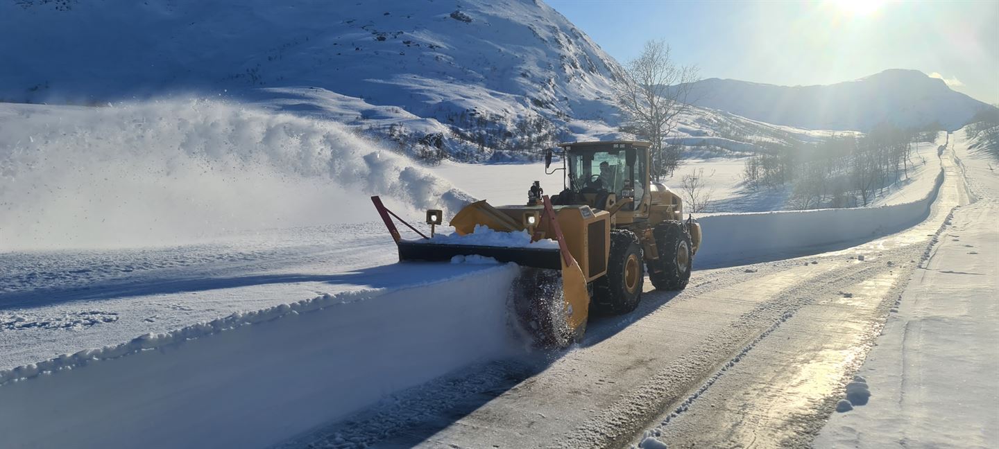 Snødekt høgfjellslandskap, Gaularfjellet, blå himmel og sol i bakgrunnen. Oransje brøytebil køyrer mot oss og brøytar snø, nybrøyta veg strekk seg bakover i bildet.