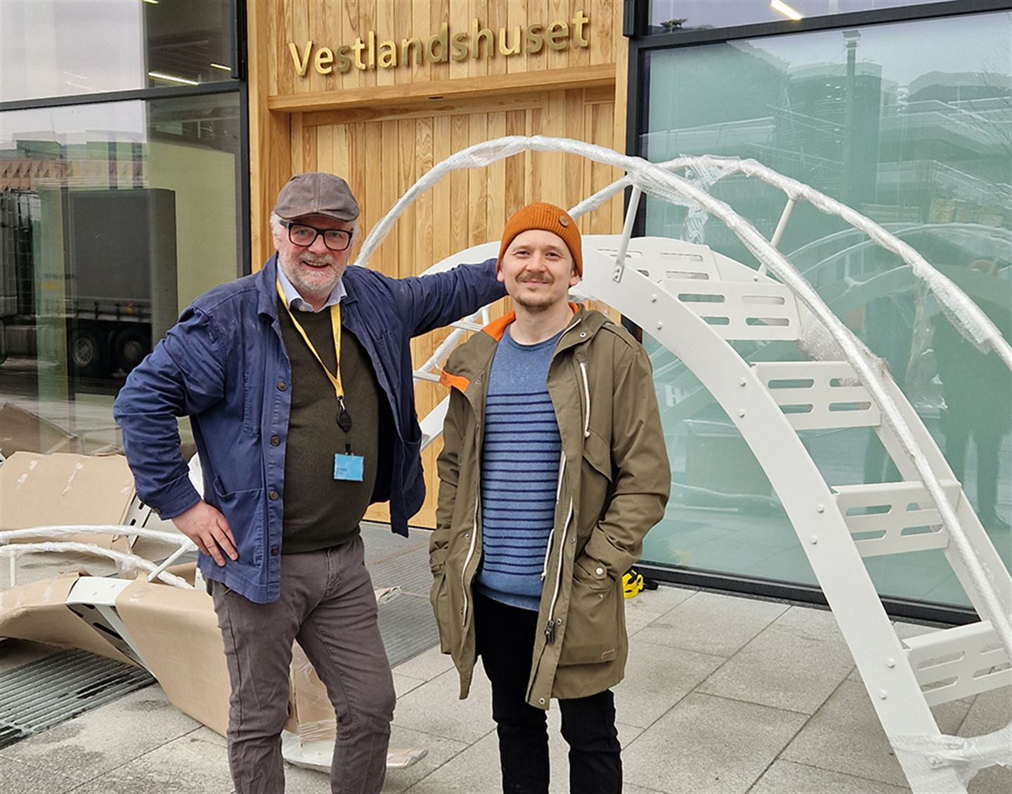 To menn utanfor framfor tredøra inn til Vestlandshuset. Det står Vestlandshuset over døra, og bak dei ligg eit kunstverk i delar.