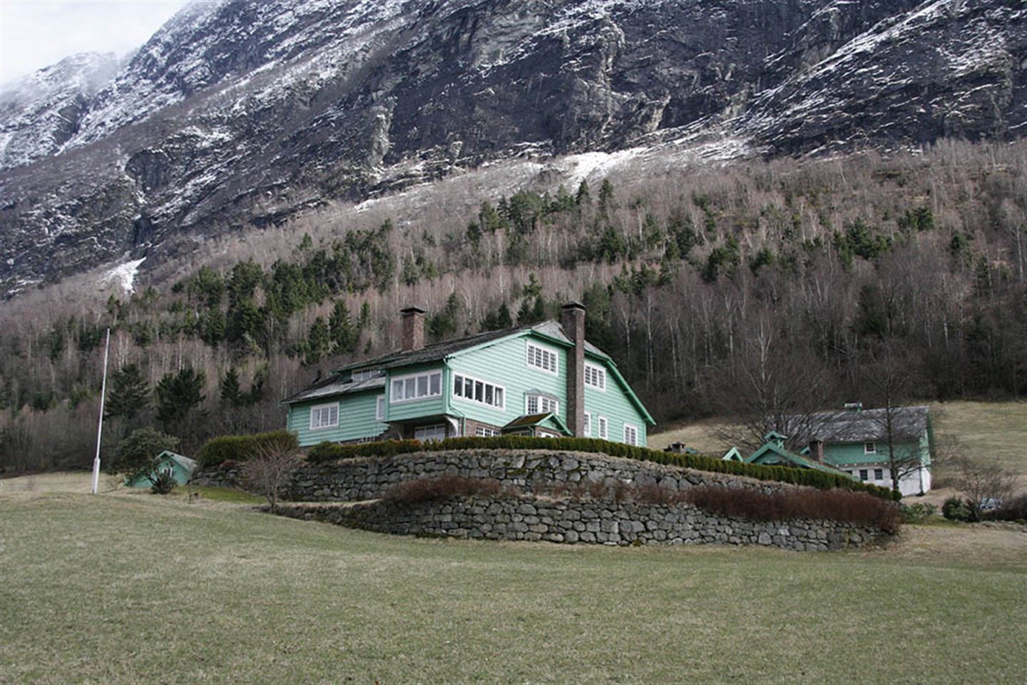 Foto av stor grønn bygning og naturen rundt med trær, fjell og 
grønn mark.