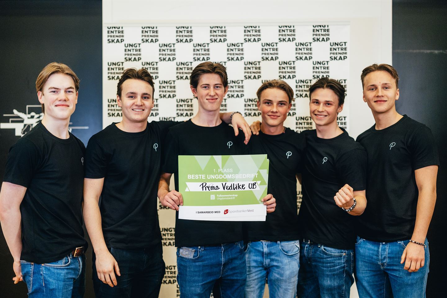 Seks unge gutar som held rundt kvarandre. Guten i midten held ein grøn plakat med teksten "1. plass Beste ungdomsbedrift".