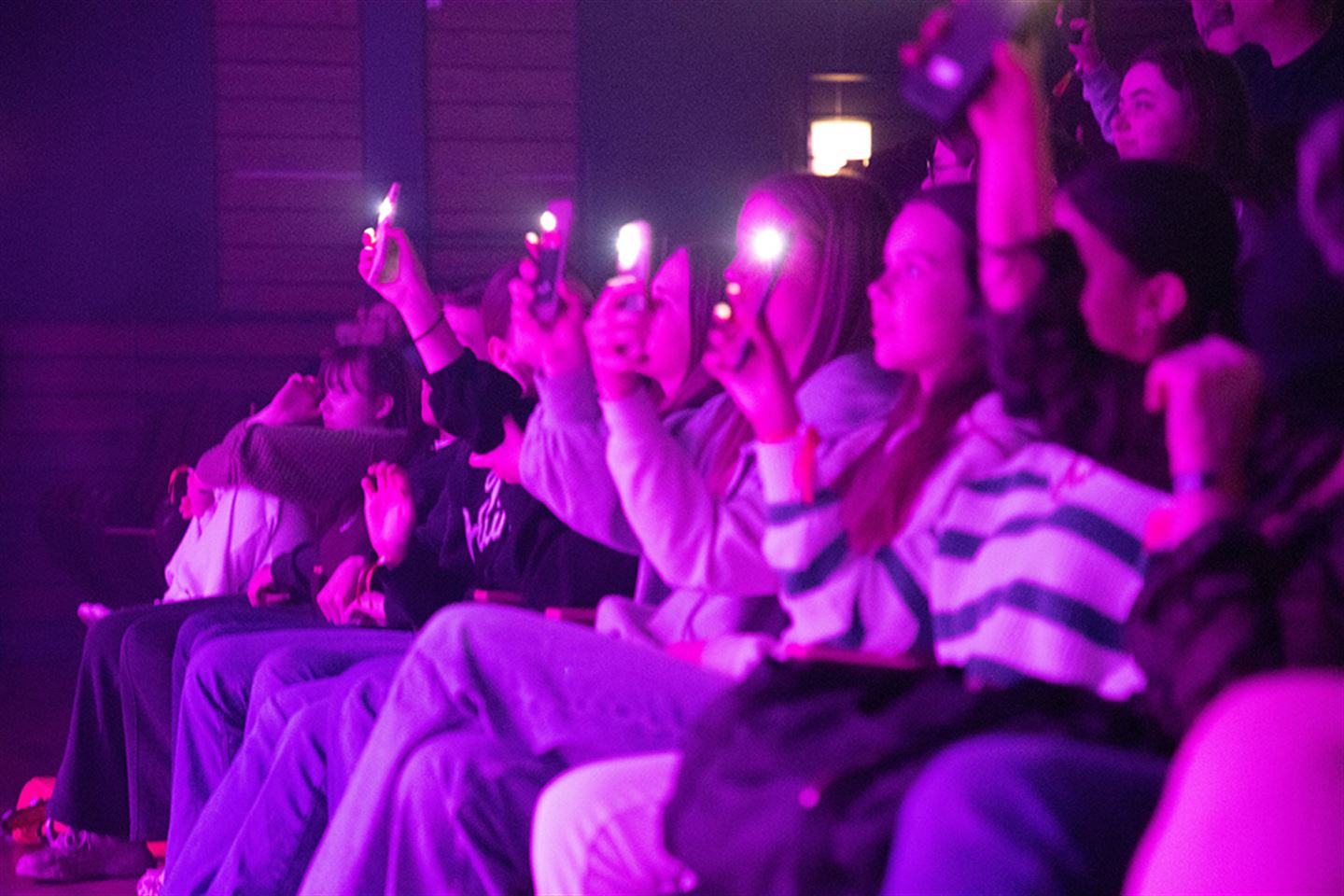 Ungdomar som held opp telefonane sine og lyser på konsert. Det er lilla lysskjær i bildet.