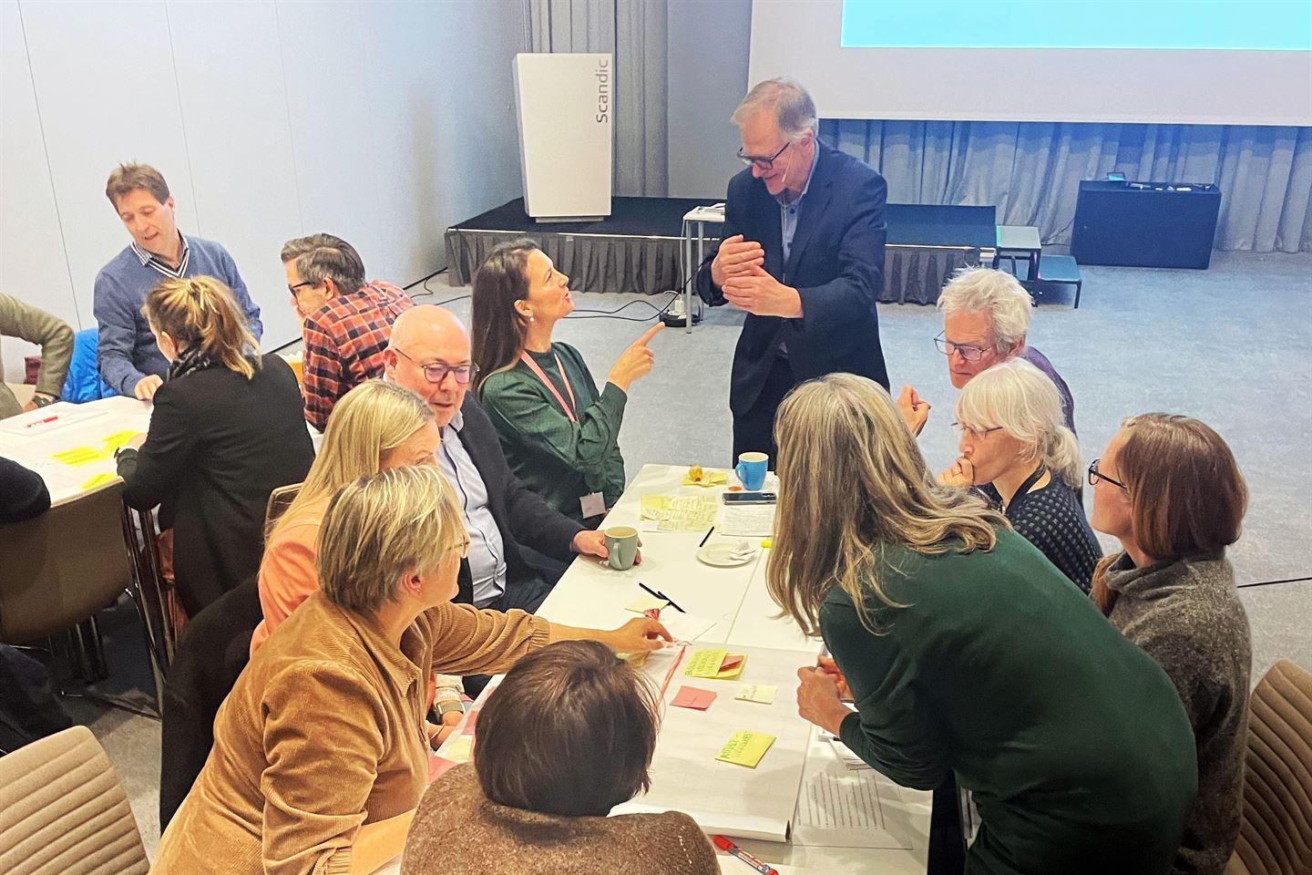 Oversiktsbilde av eit og eit halvt bord i stort lysegrått møterom, 2 menn og 7 kvinner sit rundt bordet og diskuterer, ein mann samtalar med dei ved enden av bordet. det ligg gule og rosa post-it-lappar på bordet med notatar og eit par kaffekoppar. Vi ser delvis eit bord til der det sit to menn og ei kvinne og diskuterer.