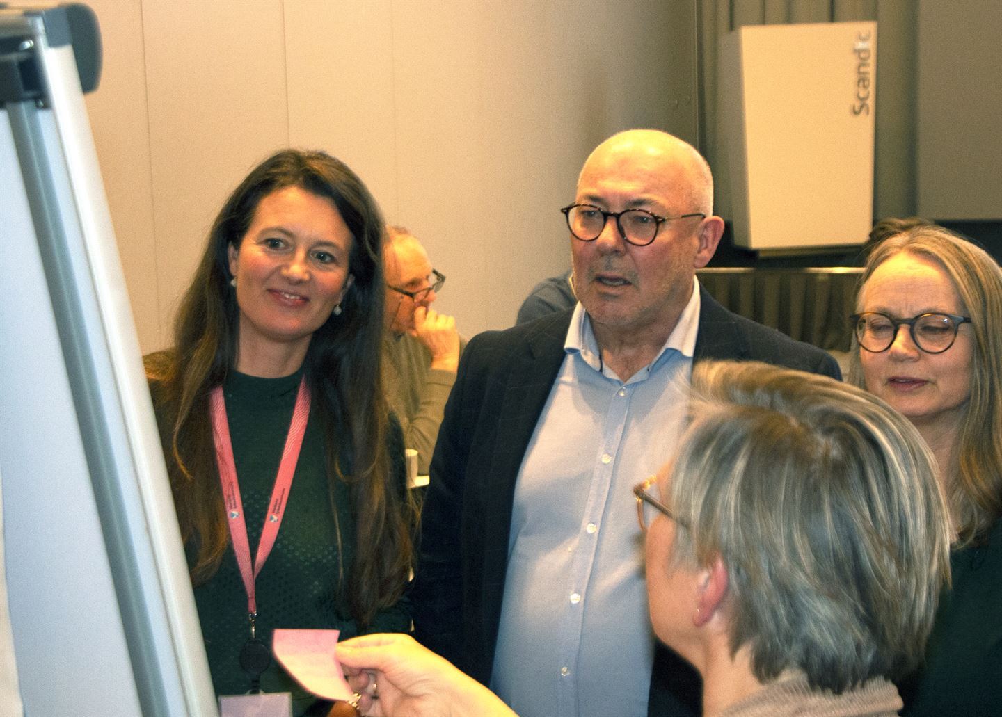 tre kvinner og ein mann står foran ei tavle med papir på, vi ser ryggen av tavla. Dei diskuterer og ei kvinne set ein rosa post-it-lapp på tavla. dei er i eit stort møterom.