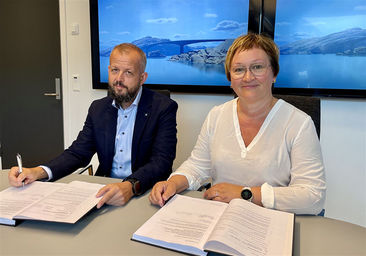 Foto av to personar som sit med dokument framføre seg på eit bord. Skjerm med illustrasjon av bru i bakgrunnen.