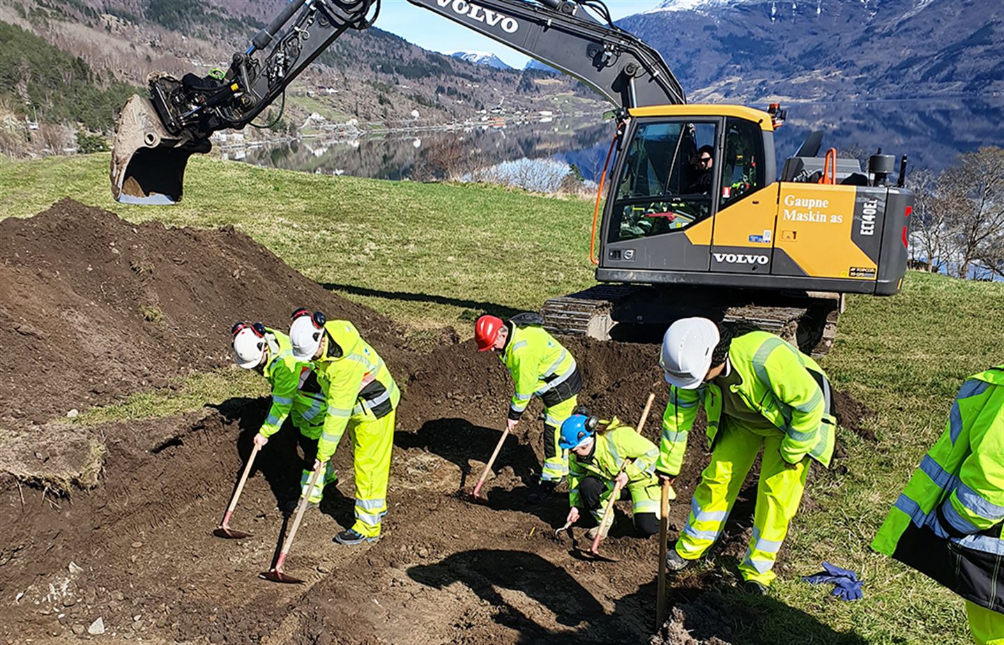 Menneske i gule arbeidskle grev i jorda, med gravemaskin i bakgrunnen.