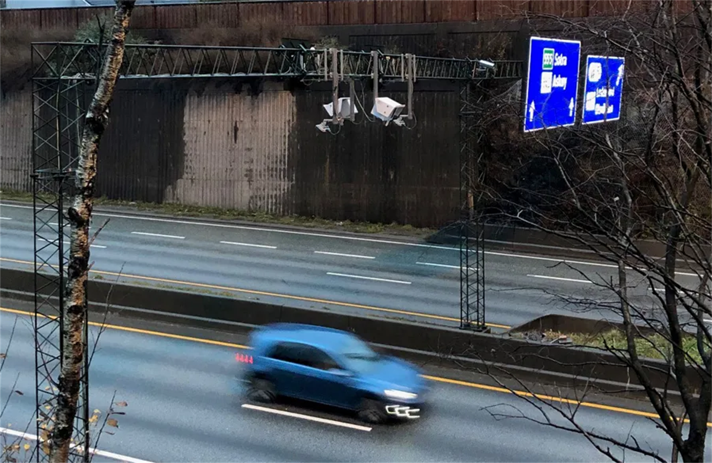 Nærbilde ovanifrå og frå sida av 4-felts grå motorveg med grå mur og brunt buskas bak, ein blå bil køyrer mot høgre, over vegbanene heng det to kamera for bompengeregistrering og to store blå skilt med tekst m.a. Sotra og Askøy. vegbanene er delt med midtrabatt og det er eit tre utan lauv i forgrunnen.