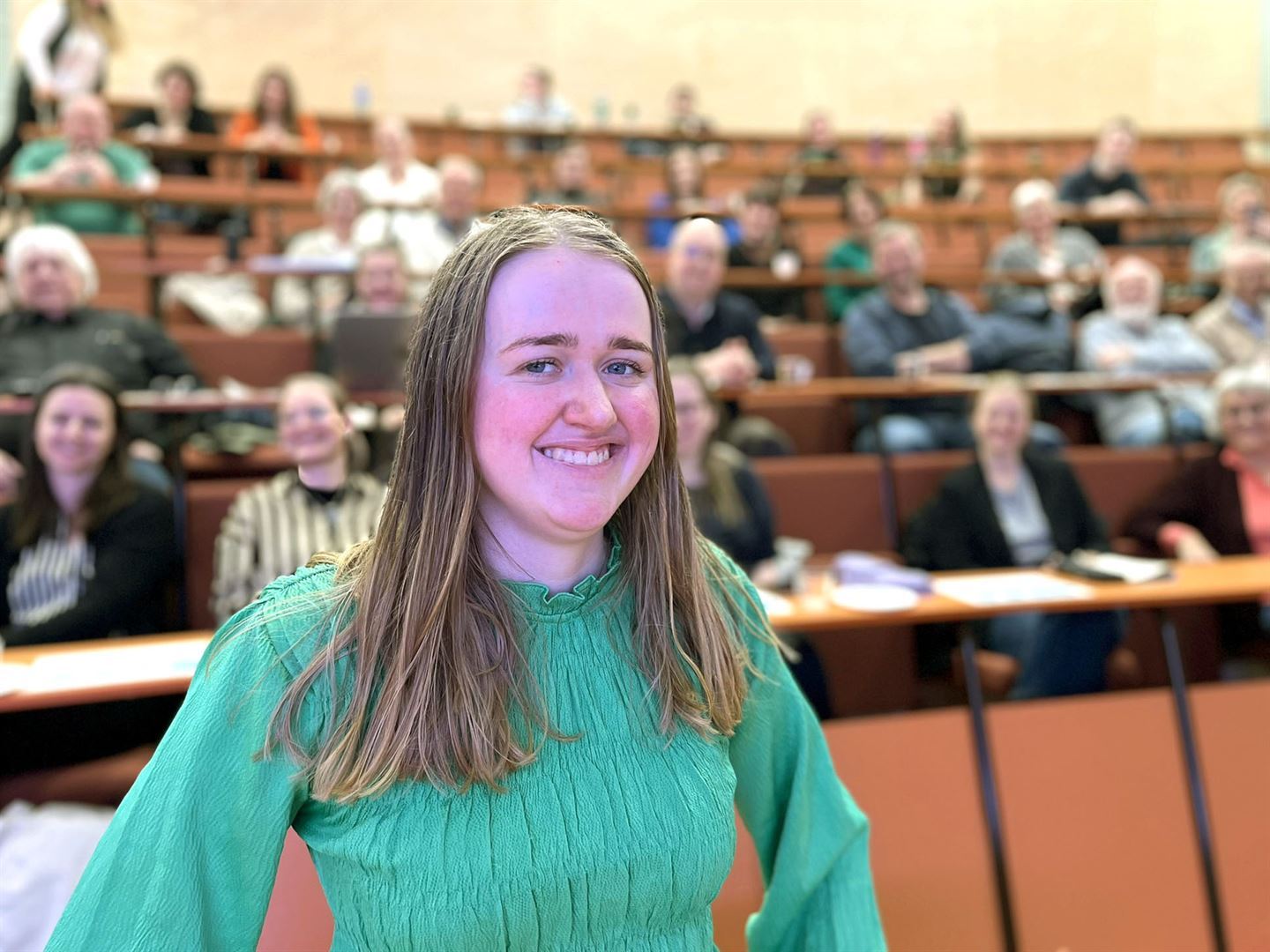 Portrettbilde av ei smilande Lisa Horpen, 22 år, i grøn bluse og langt blondt håri sal med atrium, publikum sit bak oppover seteradane i ufokus.