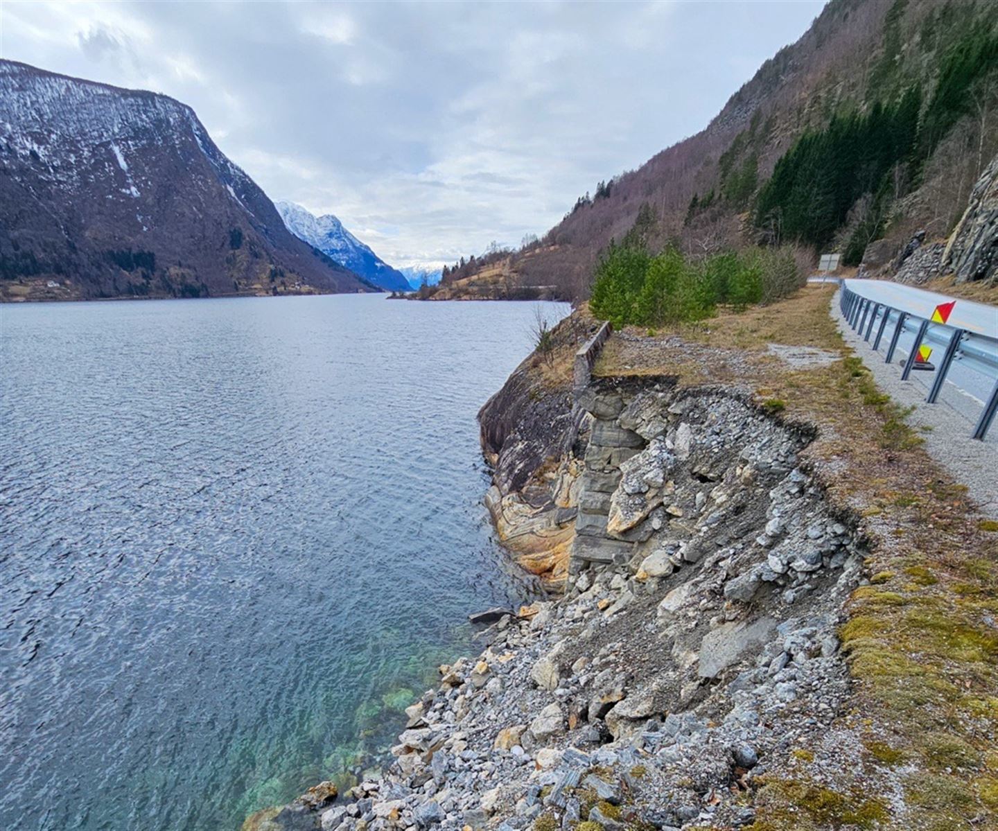 Blå fjord med fjell og veg på høgre side i bildet, muren under vegen har rasa ut i fjorden. vi ser ut fjorden mot fjell i det fjerne, vår med lauvsprett på trea og lettskya blå himmel.