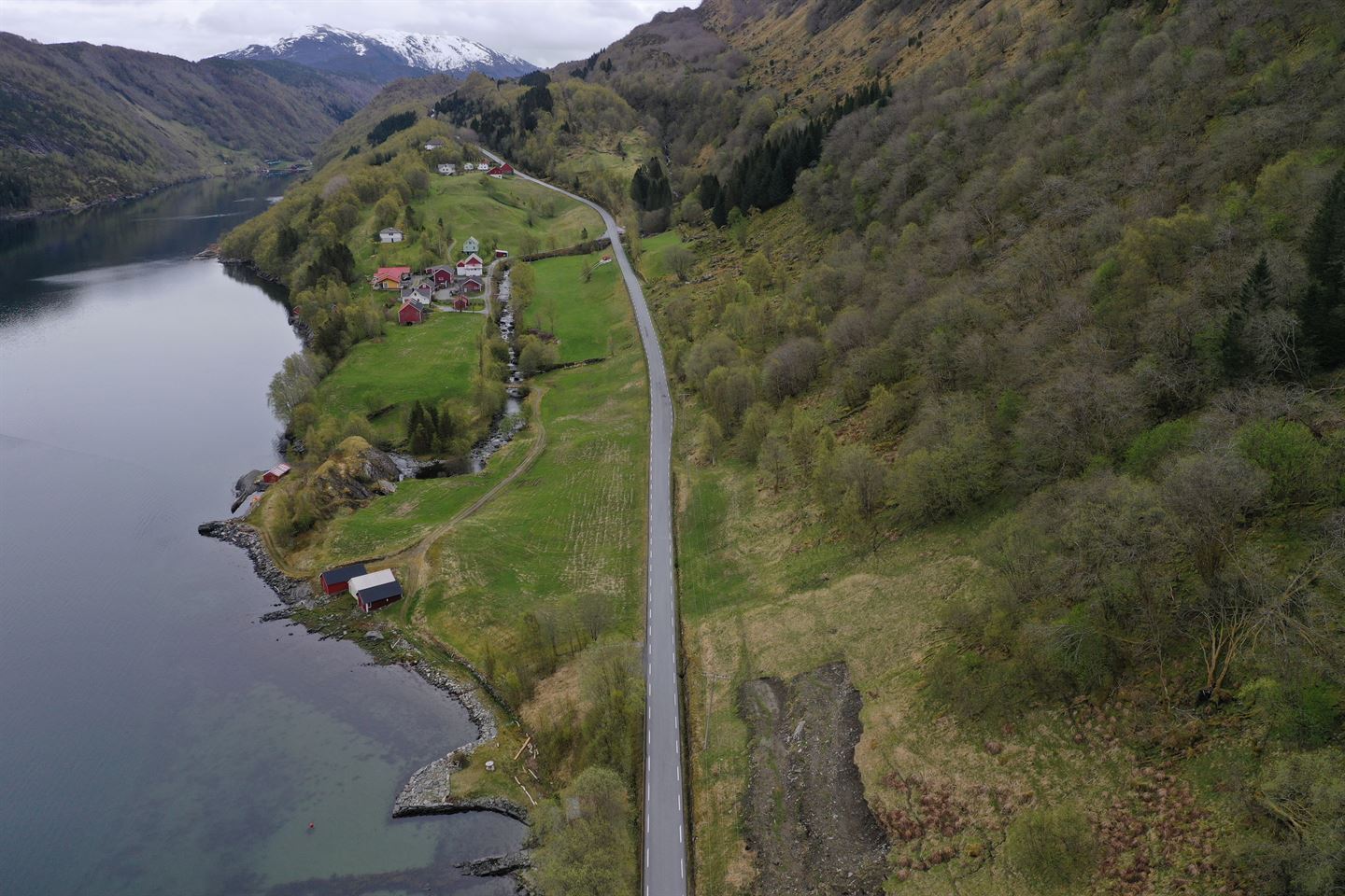 oversiktsbilde av veg med ei køyrebane som går langs fjorden, med skogkledd fjellside på andre sida. Nokre hus ligg ved vegen. Vi ser det frå lufta, det er sommar og grøne tre og grønt gras.