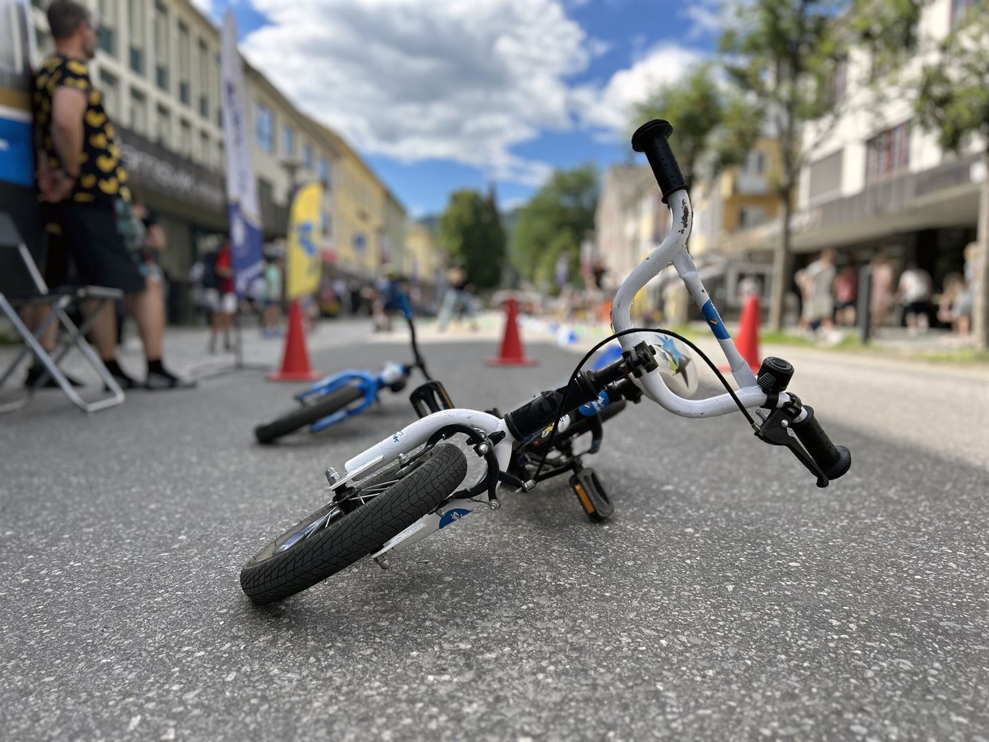 Ein barnesykkel ligg på asfalten med styret og forhjulet framst. Sykkelen er kvit og svart. Bak står tre raude kjegler.