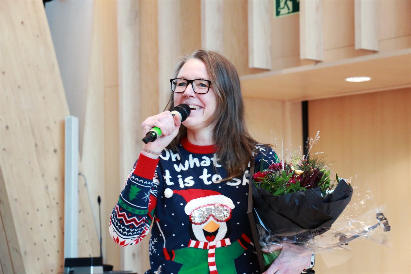 Ei smilande dame held ein mikrofon i handa. Har har mellommørkt laust hår, briller og julegenser med ein pingvin på seg. I eine armen held ho ein blomebukett.