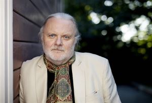 Portrettfoto av Nobel-vinnar i litteratur, Jon Fosse. Han har grått hår og grått skjegg og kvit blazer, og står lent inntil ein vegg. 