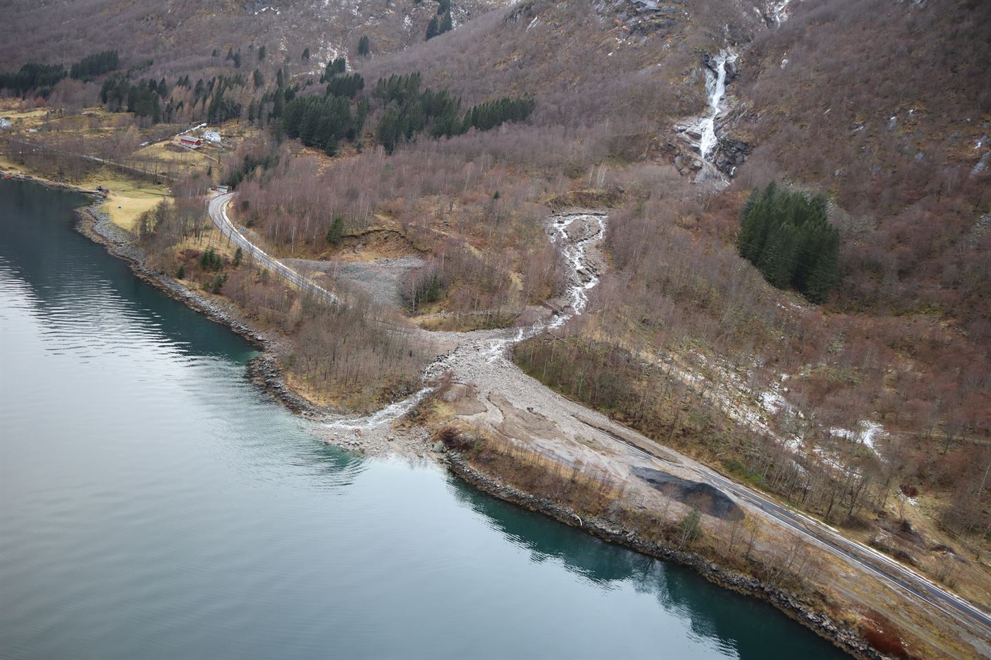 oversiktsbilde frå lufta, fjell møter fjord, veg går langs fjorden, elv kjem ned over vegen og har ført med seg stein- og jordmassar som dekker vegen. Haust, skogen har mista lauv. Gråvêr. 