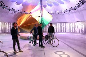 I  midten er ei solur i regnbogens farger. Den står i midten av eit rom i ein tunnel. Foran soluret står fire menn, to av dei med syklar. Mannen til venstre ser opp og held ein joystick som han styrer ein drone med. Mannen i midten held eit kamera i handa. 