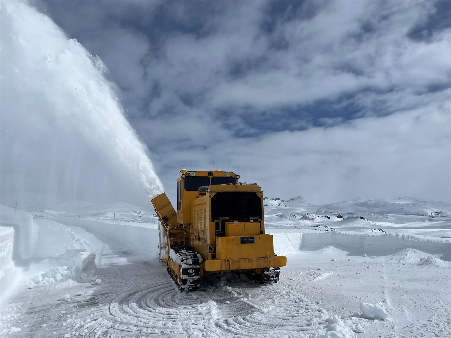 Vinterlandskap på Sognefjellet 1400 m.o.h. snødekt, snøfresemaskin i forgrunnen som brøytar og fresar snø i ei stor søyle ut over snøen til venstre, lettskya himmel og Smørstabbreene i bakgrunnen med spisse, snødekte fjell. 