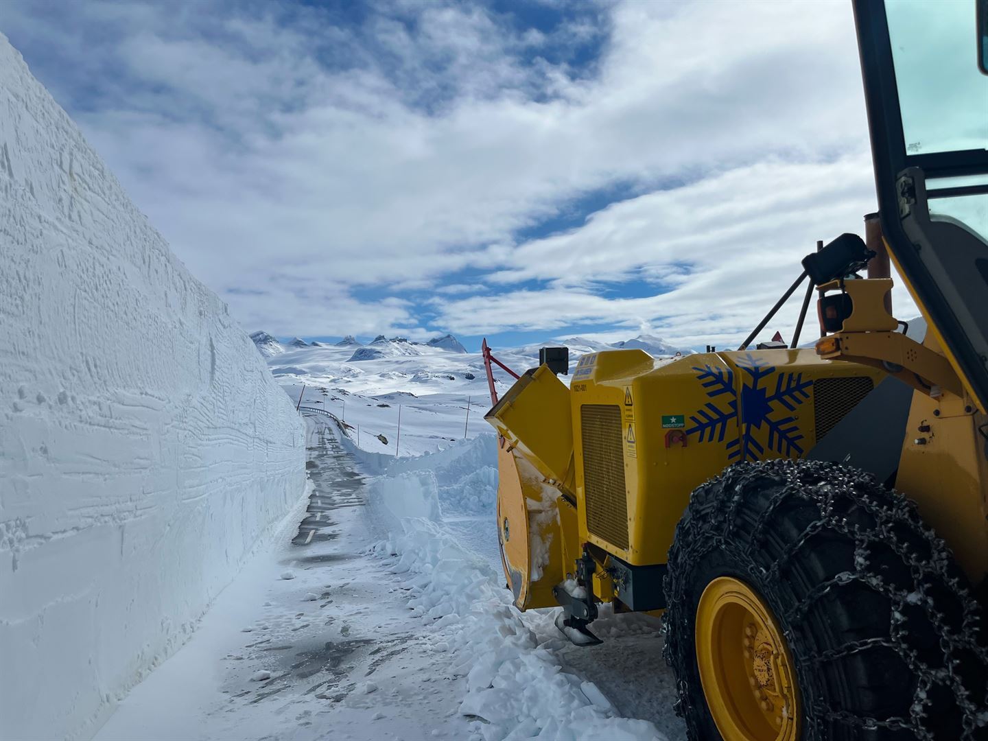 Vinterveg over høgfjellet Sognefjellet med 5 meter høg brøytekant i snø på venstre side og oransje brøytemaskin med store kjettingkledde svart hjul til venstre. Vegen strekker seg framover i bildet gjennom snøen, lettskya vêr med skyer og blå himmel, alpint fjellandskap med spisse toppar i bakgrunnen.