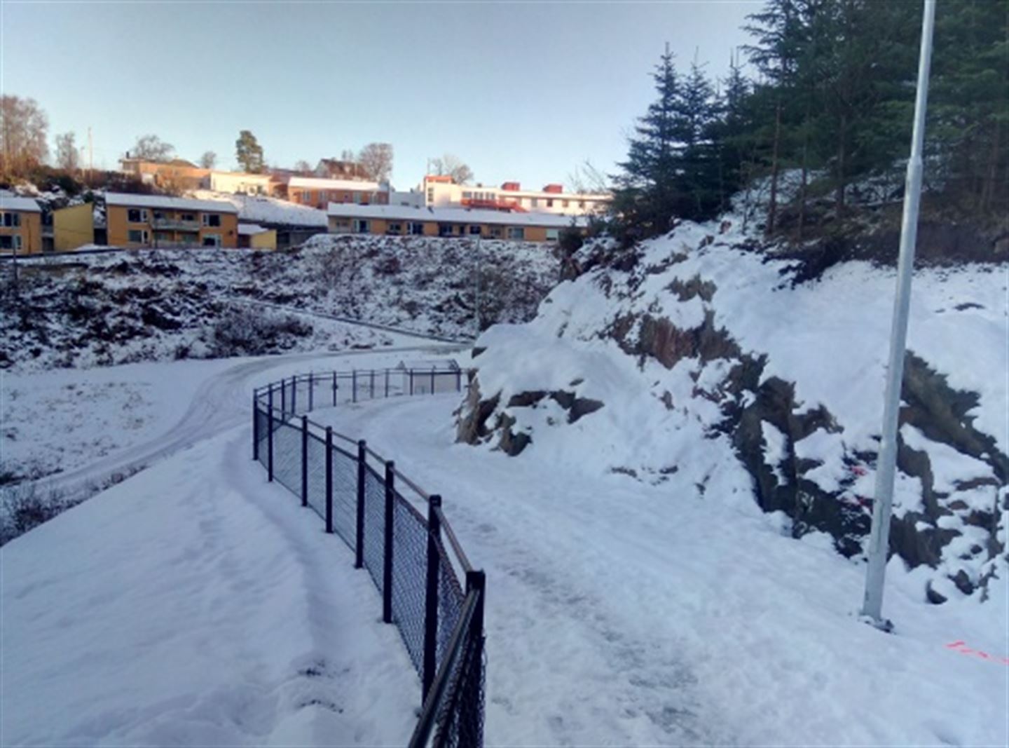 Vinter og snø, ein snødekt gangveg svingar seg opp mot eit bustadfelt, svart rekkverk og gatelys langs gang- og sykkelstien. 