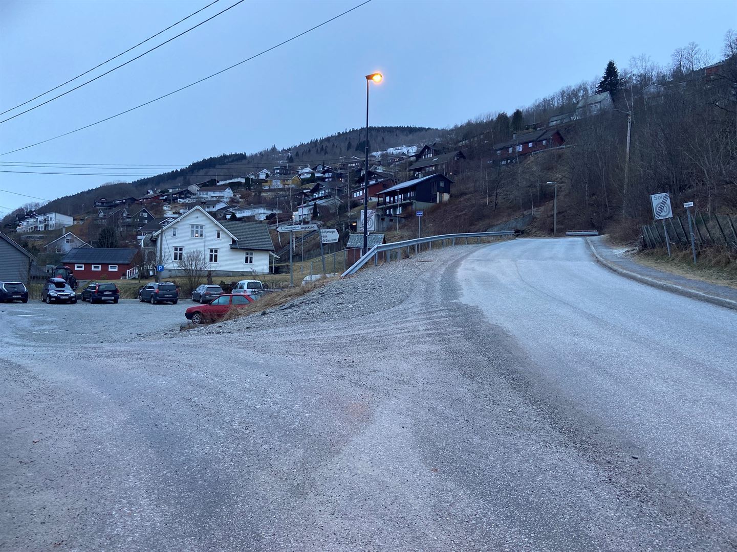 Vegkryss med grå asfalt, mykje grus, manglande fortau på venster side, gamalt stygt fortau på høgre side. 