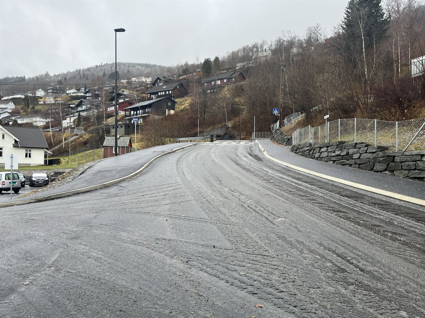 Vegkryss i regnvêr, grå asfalt, vegen går rett fram, sving til venstre i forkant av bildet. Nytt, fint fortau langs vegen på begge sider, veglys. 