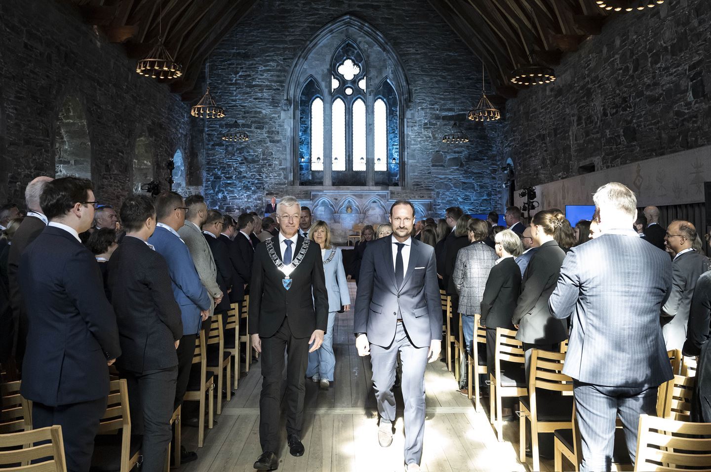 Bilde frå Håkonshallen i Bergen. To menn i dress går langs midtgangen mot kamera. På kvar side er det stoler og folk som står. Rommet er bygd i stein og det er lysekroner i taket. Foto: Thor Brødreskift.