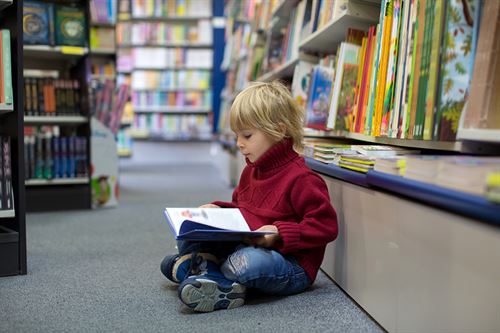 Lite barn som sit og ser i ei bok på golvet i bibliotek.