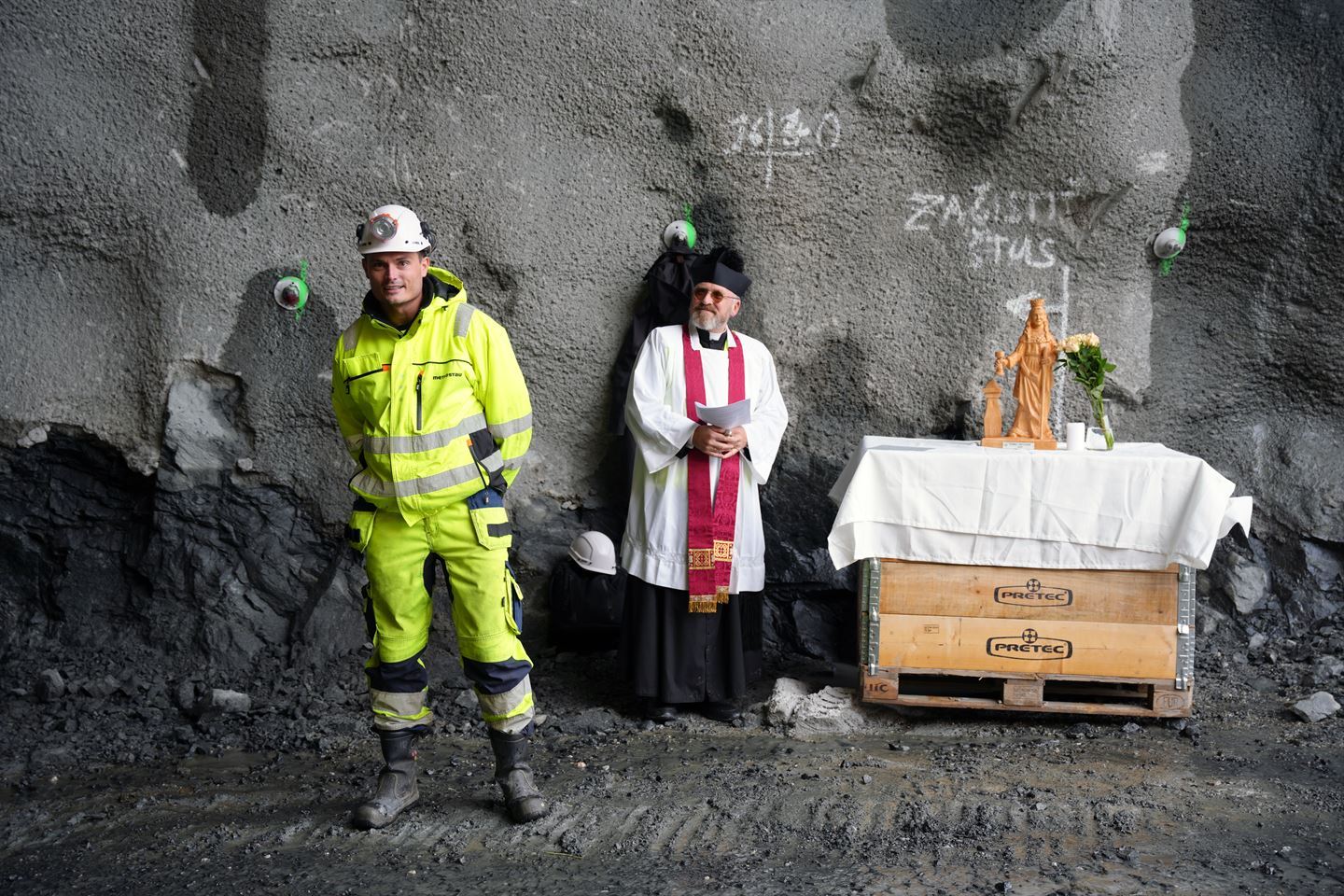 Ein mann har gul refleksbukse og jakke, kvit hjelm på hovudet. Ved sida av står ein katolsk prest med svart fotsid kappe, kvit skjorte og raudt band over skuldra, på hovudet har han ein svart, liten firkanta hatt. Ved sida av dei to er eit alter med kvit duk. Oppå duken står ei trestatue av ei kvinne og ein vase med kvite roser. Foto: Silje Alvsaker/Vestland fylkeskommune 