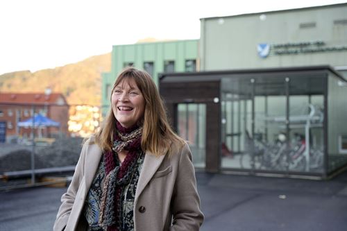 Ei dame med brunt hår, grå kåpe og sjal i halsen. Bak ho er eit glasskur med syklar og ein skulebygning. 