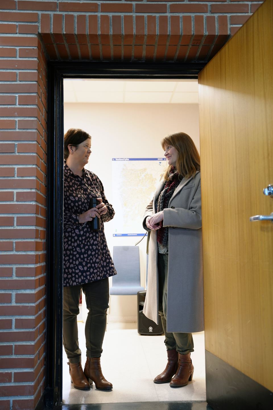 To damer står i ei døropning og snakkar saman. Ei dame har mørkt kort hår, bluse med prikker, mørk bukse og brune skinnstøvlettar. Dama ho prater med har mørkt langt hår, grå kåpe, mørk bukse og brune skinnstøvlettar. 