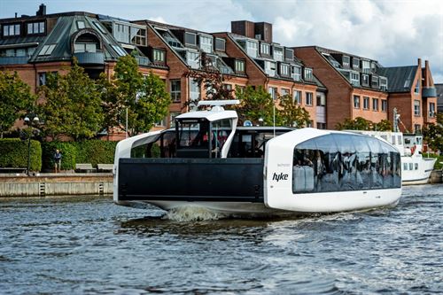 El-båt i fart med strandpromenade og hus i bakgrunnen.