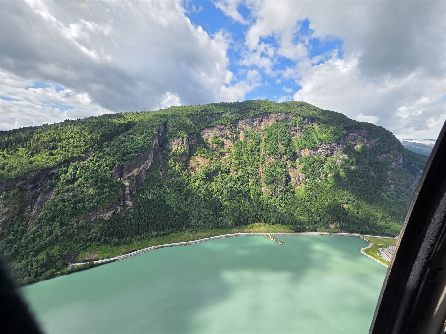 bilde av fjellside med grønn skog, blå himmel, blå fjord i forkant av bildet, ein veg går langs strandlinja. Fjellsida har opne parti med berg.