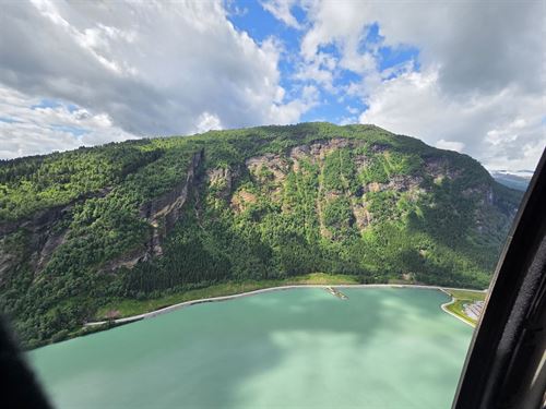 bilde av fjellside med grønn skog, blå himmel, blå fjord i forkant av bildet, ein veg går langs strandlinja. Fjellsida har opne parti med berg. 