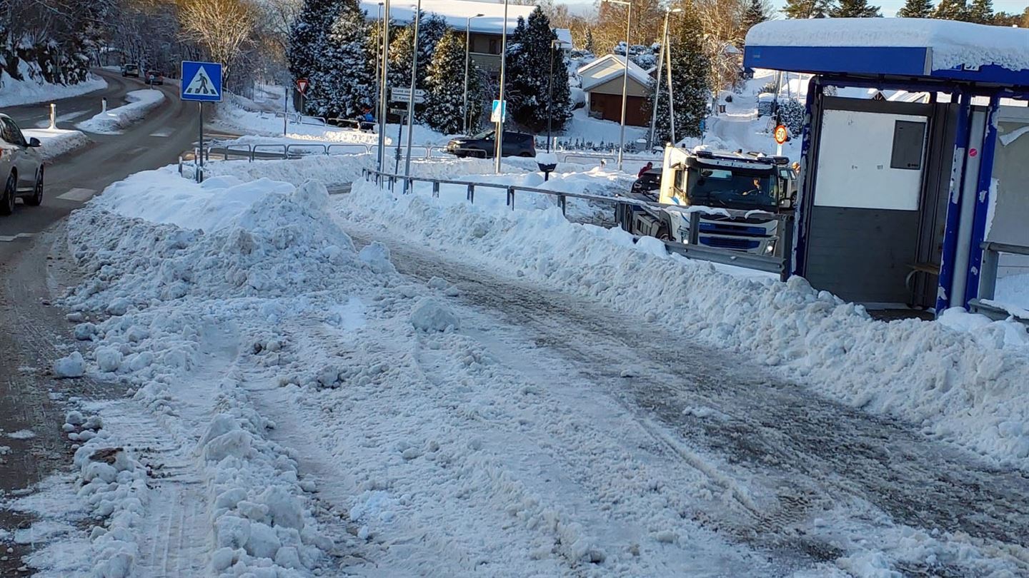 veg med grå asfalt, bil køyrer frå oss. På gang- og sykkelvegen til høgre for vegen er det mykje snø, delvis brøyta vekk, med ei busslomme der snøen har hopa seg opp i brøytekantar. Vi ser ein parkeringsplass bak busslomma, blå himmel og snødekt landskap bak.