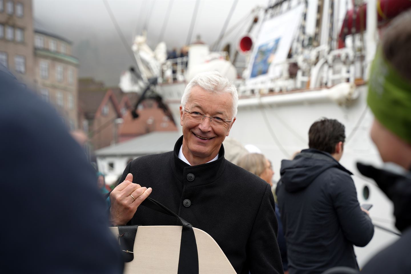 Ein smilande mann med kvitt hår, briller og svart frakk held ei veske i handa. Bak han ligg eit stort skip til kai. Foto: Silje Alvsaker/Vestland fylkeskommune