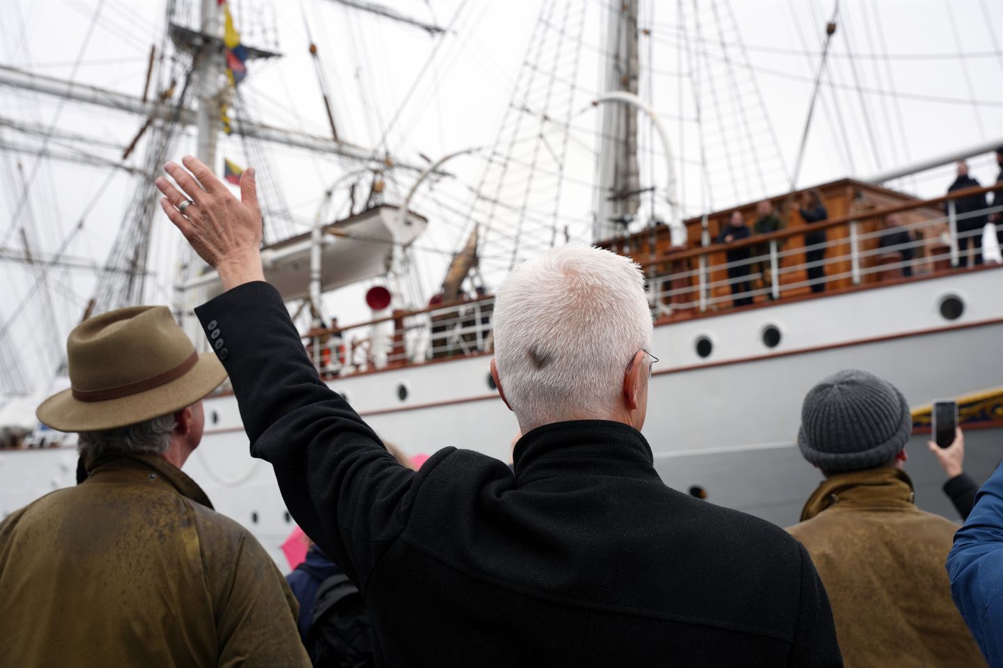 Ein mann med kvitt hår og svart frakk vinkar til ei skute som er i ferd med å legge frå kai. Foto: Silje Alvsaker/Vestland fylkeskommune