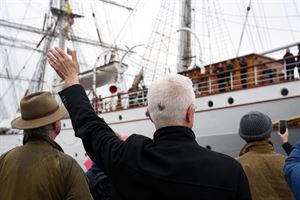 Ein mann med kvitt hår og svart frakk vinkar til ei skute som er i ferd med å legge frå kai. Foto: Silje Alvsaker/Vestland fylkeskommune