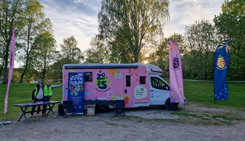 Rosa bubil med profilering for friluftslivets år 2025 og to menneske og fleire bannerflagg utanfor.