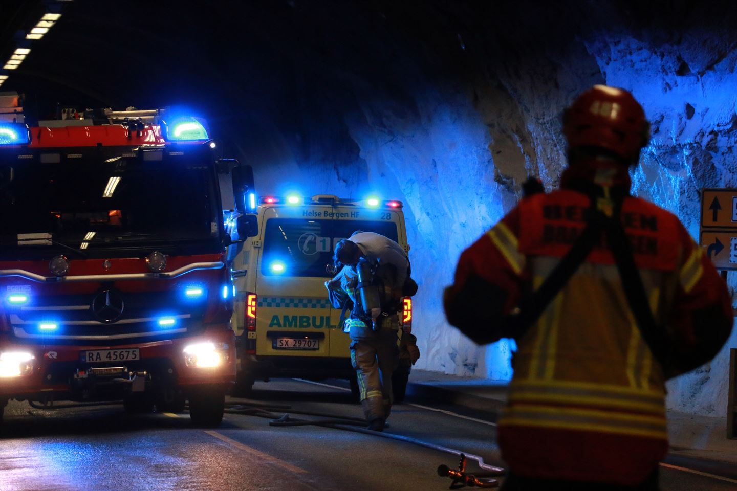 Ein raud brannbil med blålys står parkert i ein tunell. Ved sida av står ein gul og grøn ambulanse, denne også med blålys på. Ein brannkonstabel går mot ambulansen med ein skada pasient hengande over skuldra. I forgrunnen står ein brannkonstabel med gul og oransje jakke og raud hjelm med ryggen mot kamera. Foto: Silje Alvsaker