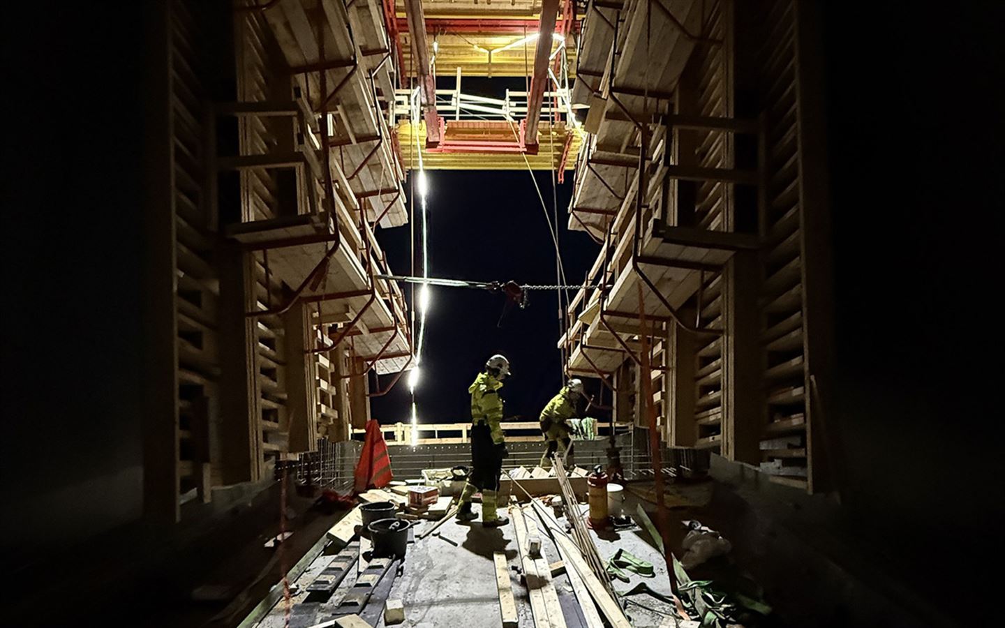 Vi ser inn i eit mørkt utandørs arbeidsrom i betong med forskalingar av tre på veggar og tak, stor takhøgde, to arbeidarar står i rommet som er ope ut mot nattehimmel bak dei. Arbeidarane har gult arbeidstøy og kvite hjelmar, og plankar i tre ligg på betonggolvet. Ein enkelt lyssøyle lyser opp området der dei jobbar, det andre ligg i mørket. 