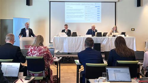 Stort rom med lyse veggar og tak, fleire rekker med bord og stolar i forkant der vi ser menn og kvinner sitte ved pultane med PC på bord framfor seg med ryggen mot kamera. Foran rekkene med desse fylkespolitikarane er det eit bord der fylkesdirektør Robert Rastad, fylkesordførar Jon Askeland og ei kvinne som er utvalssekretær sit med andletet mot rekkene med folk. til venstre for dette dirigentbordet står det ein talarstol, der ein fylkespolitkar er i gang med sitt verbale innlegg. 