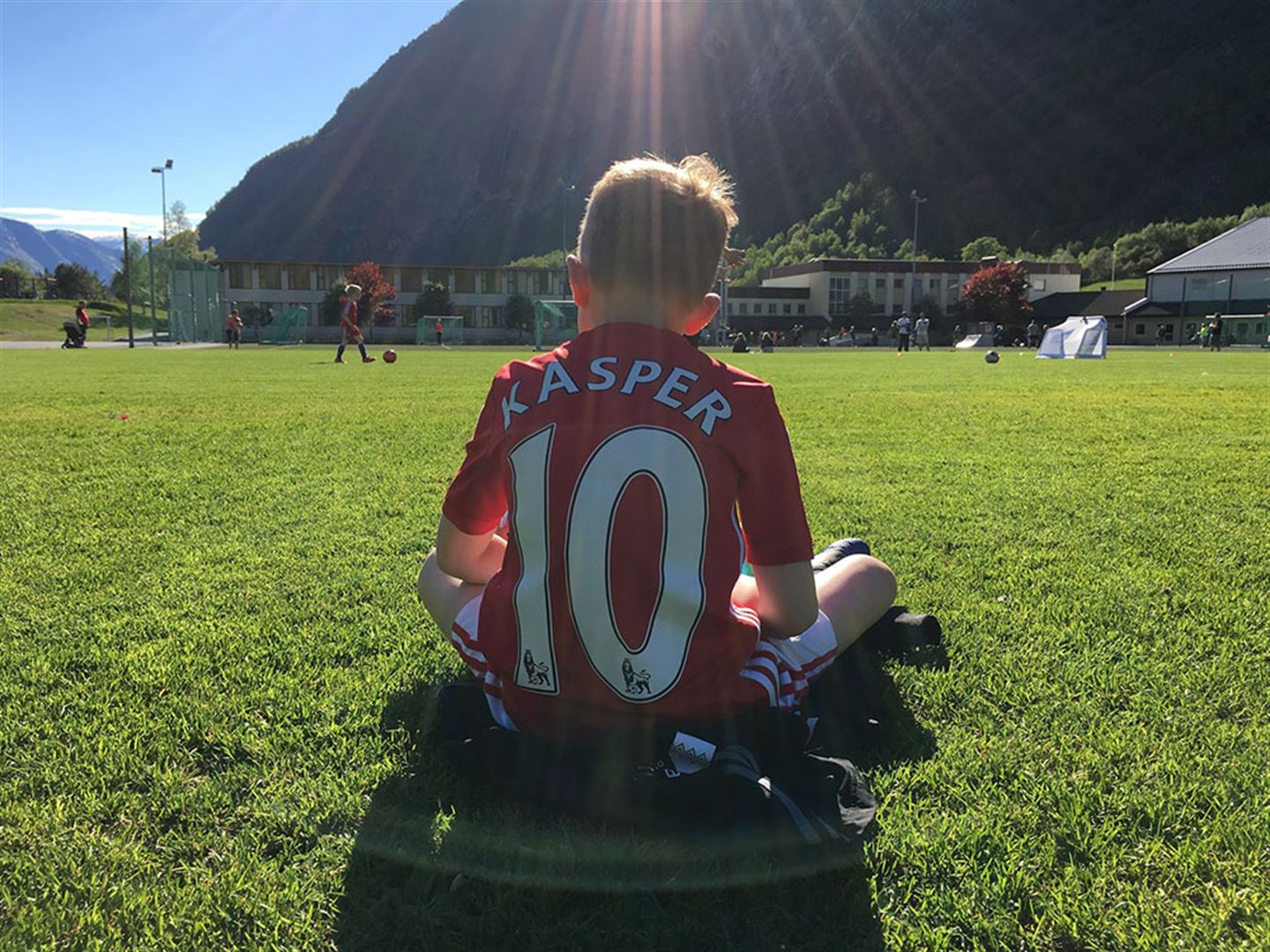 Gut som sit på fotballbane med ryggen til kameraet. På ryggen står det talet 10 og Kasper. I bakgrunnen er det ungar som spelar fotball.