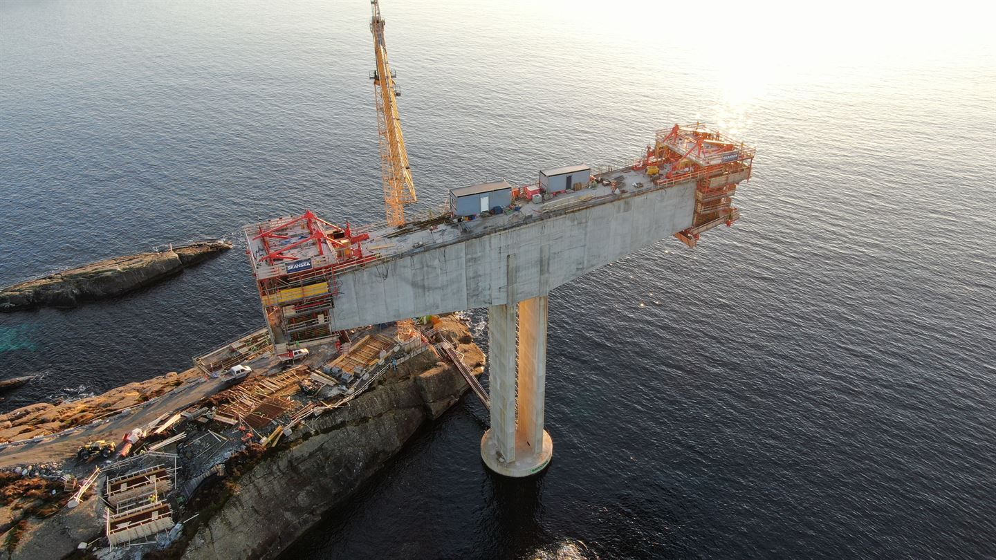 Bilde er tatt ovanfrå frå oppe i lufta. Vi ser ned på sjø og ein holme som stikk ut i sjøen, sola skin på sjøen øvst i høgre hjørne. Rett ved holmen stig ei betongsøyle opp av havet. oppå søyla blir det jobba med maskiner og mannskap, på kvar ende av toppen av søyla er det ein konstruksjon der brudekket blir støypt meter på meter. På holmen står det ei gul heisekran, og det er ein anleggsveg og tegn til byggeakvititet på holmen. 