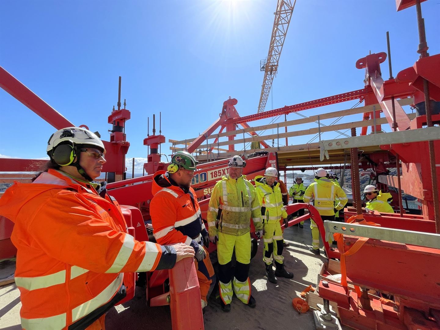 To menn i sterkt oransje arbeidstøy med kvite hjelmar og fem arbeidsmenn i gult arbeidstøy m kvite hjelmar står på betongdekke oppe på ei søyle ute i havet blant holmar og skjer. Blå himmel. Dei er omgitt av stålbjelkar og konstruksjojnar i sterkt oransen, og ei gul heisekran heng over dei. 