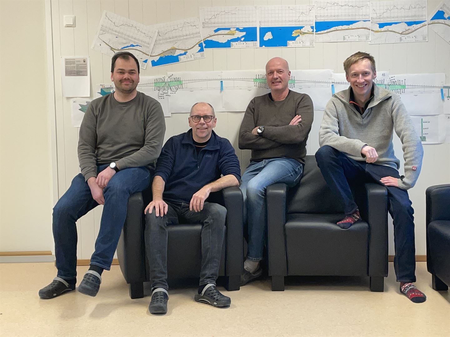 Gruppebilde av 4 personar som sit på armlenene og i setet på to svarte lenestolar i skinn opp mot ein lys vegg, iført fritidsklede: jeans og gensarar i blå- og gråtonar, og i crocs-sko eller sokkelesten. Alle er menn og alle smiler til kamera. På veggen bak dei er det hengt opp planteikningar av det som skal bli Ytre Steinsund bru, i blå og brune fargar. 